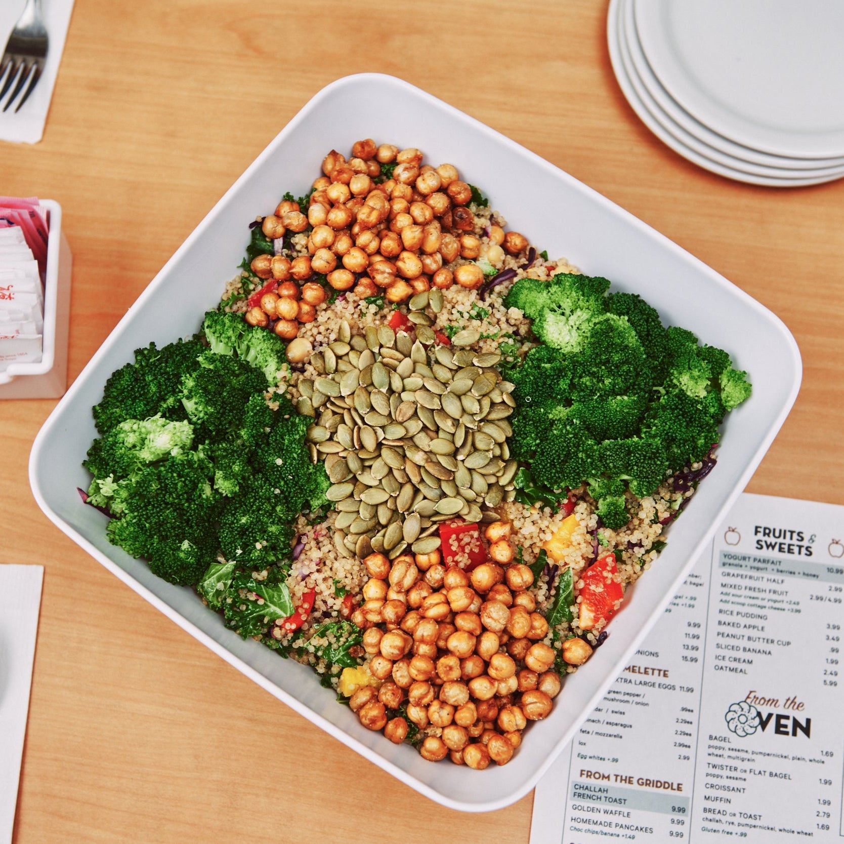 Plated salad with chickpeas, greens, and seeds on a wooden table with a menu and cutlery.