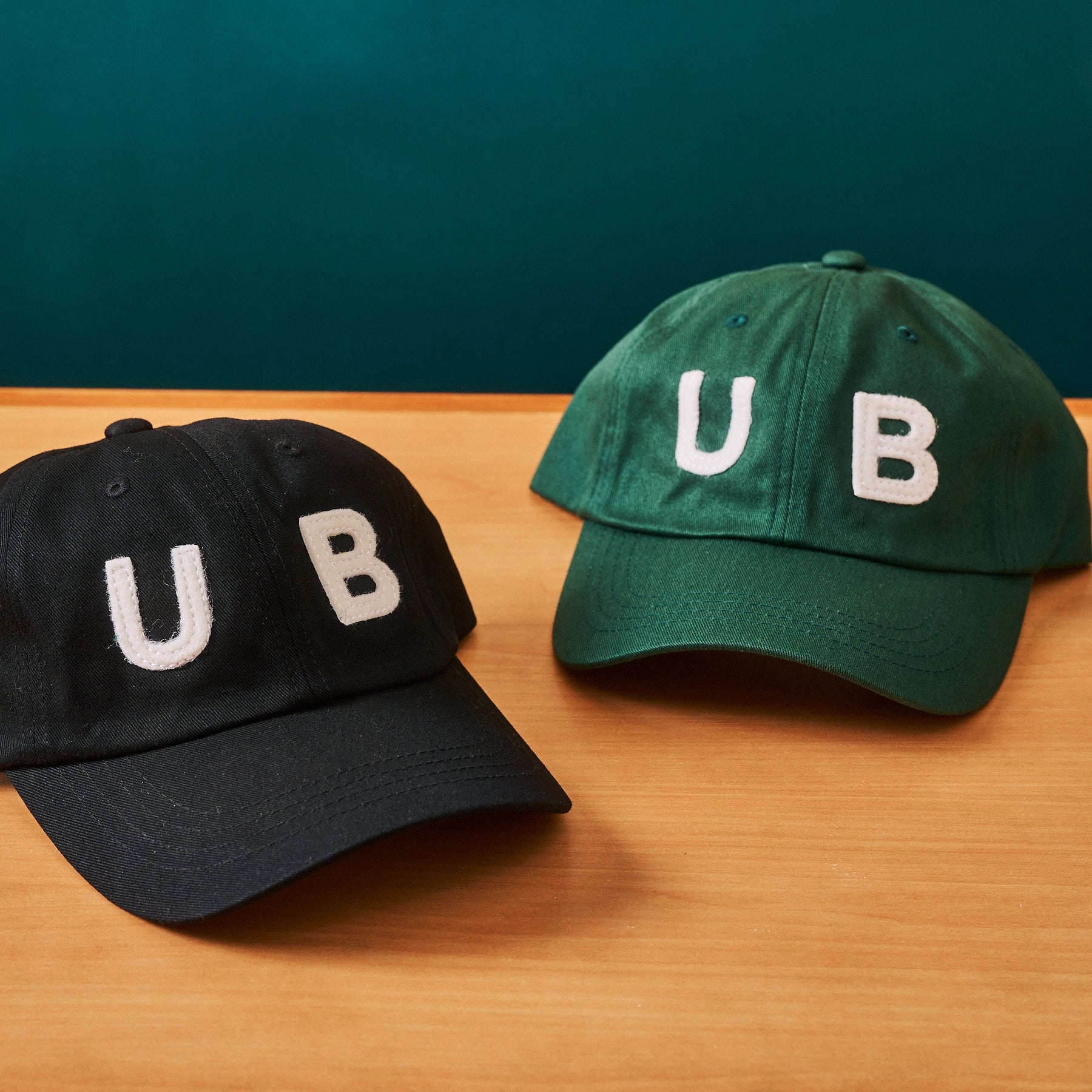 Two baseball caps with 'UB' on a wooden surface and teal background