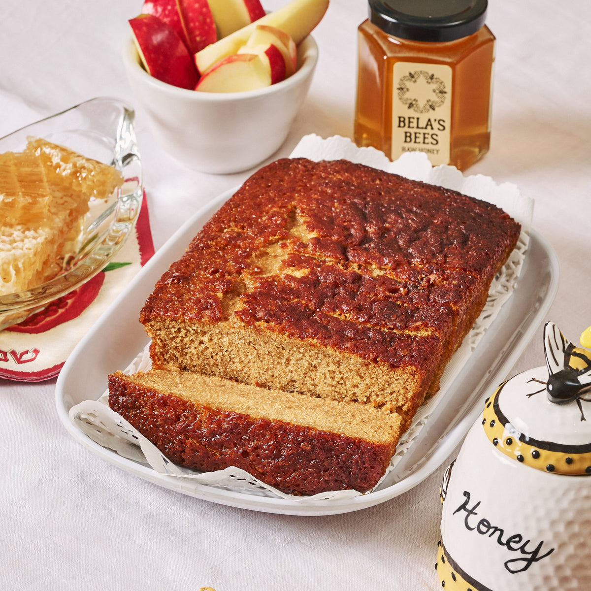 Loaf of bread on a plate with a jar of honey and a bowl of apples in the background.