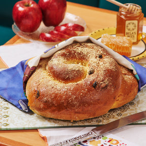 A round challah bread with raisins, partially covered with a white cloth, displayed on a table with apples and honey in the background, suggesting a setting for Rosh Hashanah celebrations.
