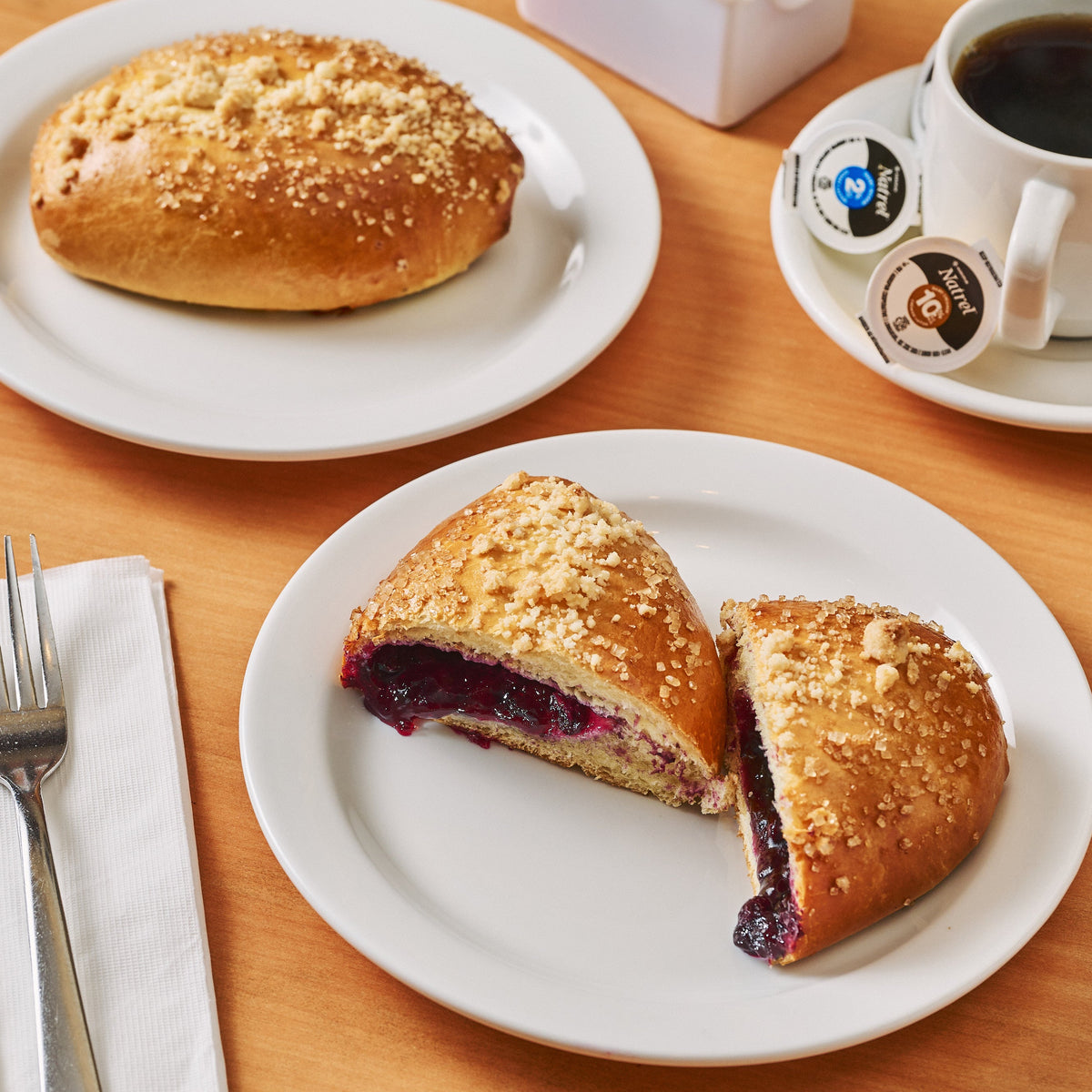 Blueberry Bun on a plate with a cup of coffee and sugar packets on a wooden table.